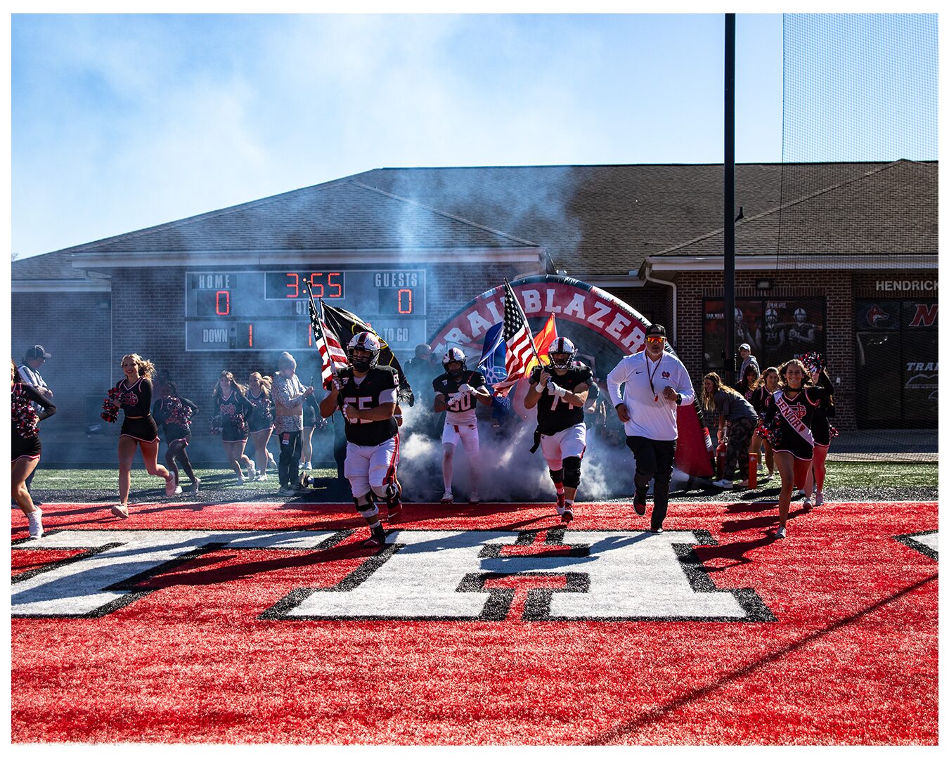 The Trailblazers run out through a smoke tunnel onto the NGU field under a clear sky as cheerleaders raise flags.